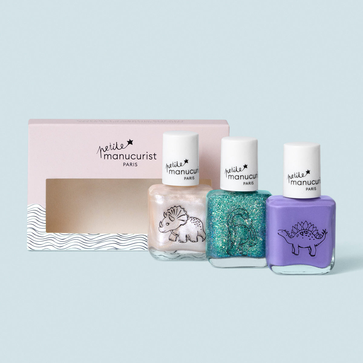 Trois flacons de la collection Wild Life de Manucurist — blanc nacré avec un tricératops, bleu sarcelle pailleté avec un brontosaure et violet avec un stégosaure — sont présentés devant une boîte rose ondulée « petite manucurist Paris » .