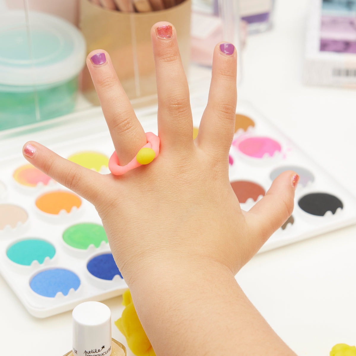La main d’un enfant aux ongles colorés utilisant le Trio Betty Scarlett Stella de Manucurist est présentée au-dessus d’une palette de peintures aquarelle, de fournitures artistiques et d’une bague rose et jaune dans un espace de travail lumineux et créatif.