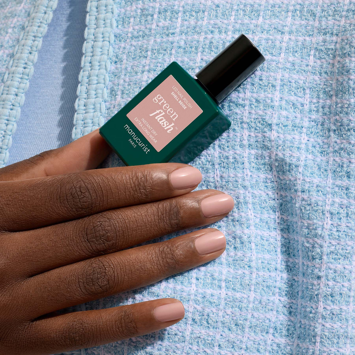 A hand with neatly manicured, Shell Beige nails holds a Manucurist Green Flash gel polish bottle against a textured light blue fabric background.