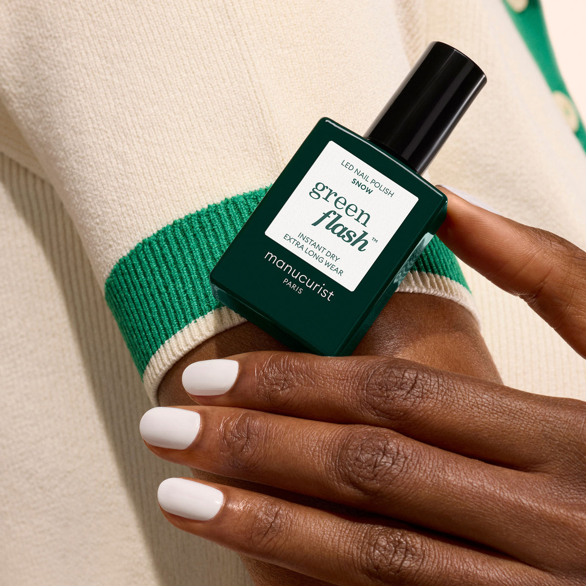 Une main portant le vernis blanc pur "Snow" de Manucurist tient un flacon vert foncé de vernis gel Manucurist Green Flash contre un pull crème bordé de vert.
