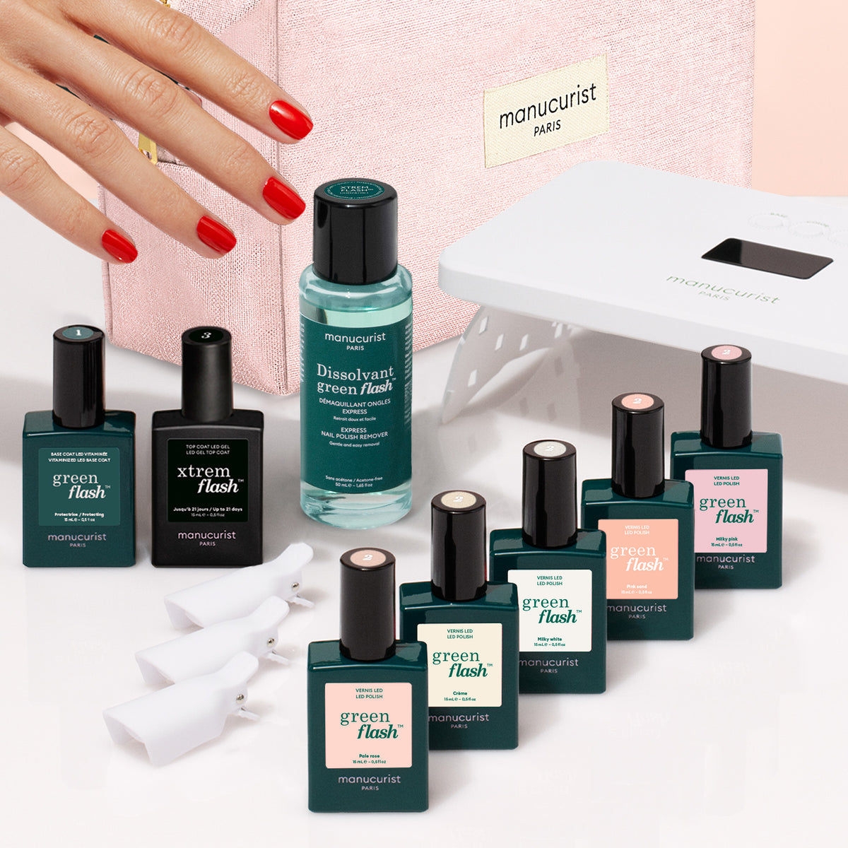 Une main aux ongles vernis de rouge se tend vers un Super Starter Kit de vernis semi-permanent LED Soap Nails, avec des flacons, du dissolvant, une lampe LED et des pinces disposés devant une trousse de maquillage rose pour réaliser une manucure ultra-brillante.