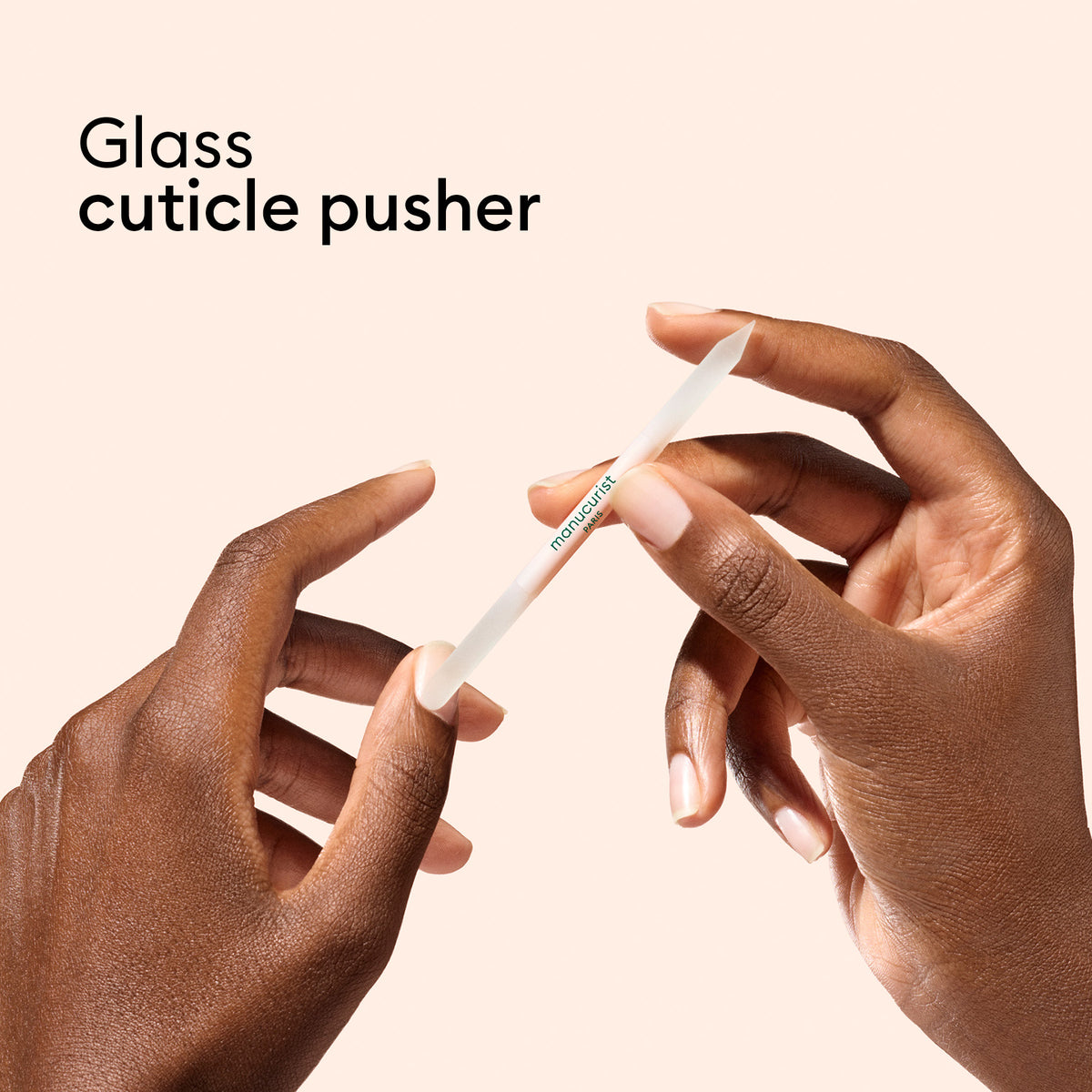 Deux mains tiennent un repousse-cuticules en verre sur un fond pêche. Le texte indique « Trio d’accessoires pour ongles en verre » par Manucurist-us. L’outil repousse délicatement les cuticules — idéal à associer à votre lime à ongles en verre préférée pour des ongles impeccables.