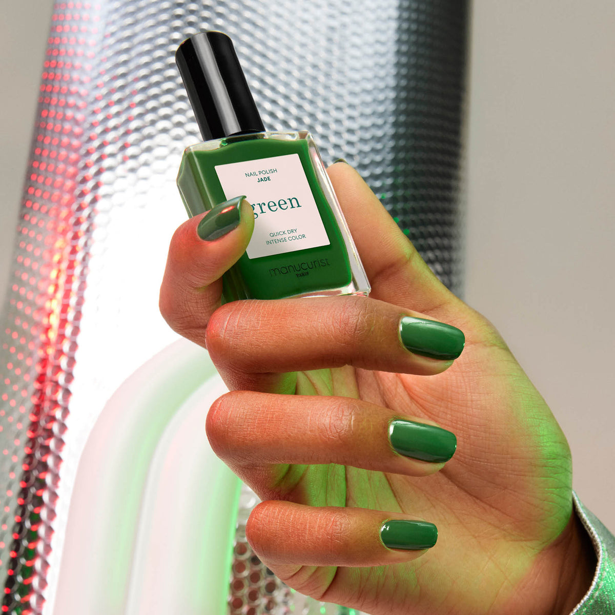 Une main aux ongles soigneusement manucurés et peints en "Jade" par Manucurist, une teinte vert minéral aux sous-tons froids, tient le flacon de vernis assorti sur un fond texturé métallique aux reflets de lumière verts et rouges.