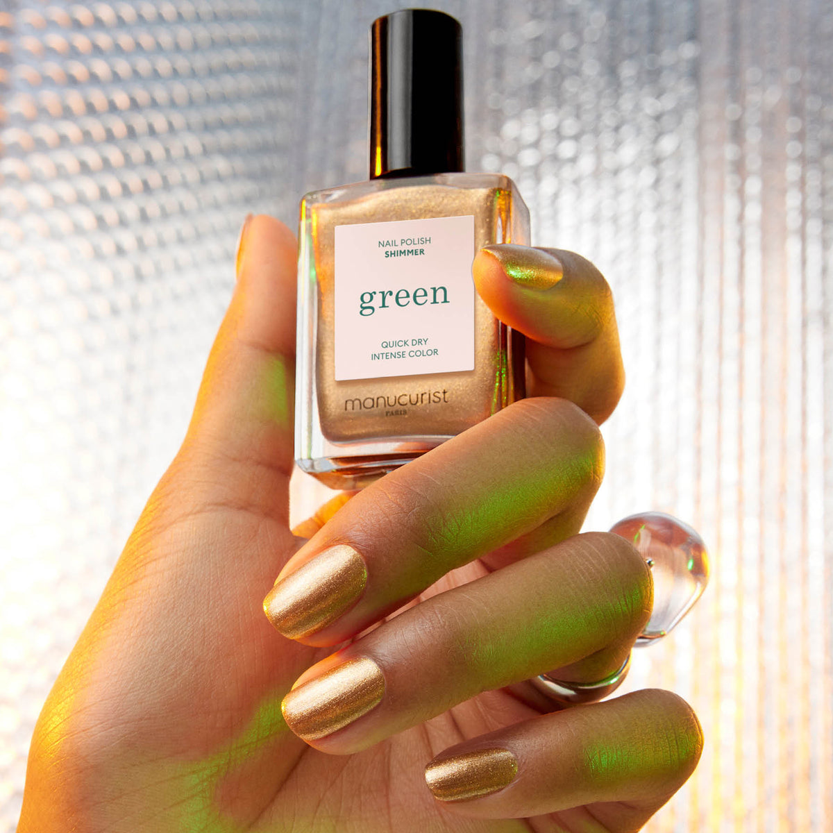 A hand with shimmering metallic gold nail polish from Manucurist holds a matching "Shimmer" bottle by Manucurist against a textured, softly lit background.