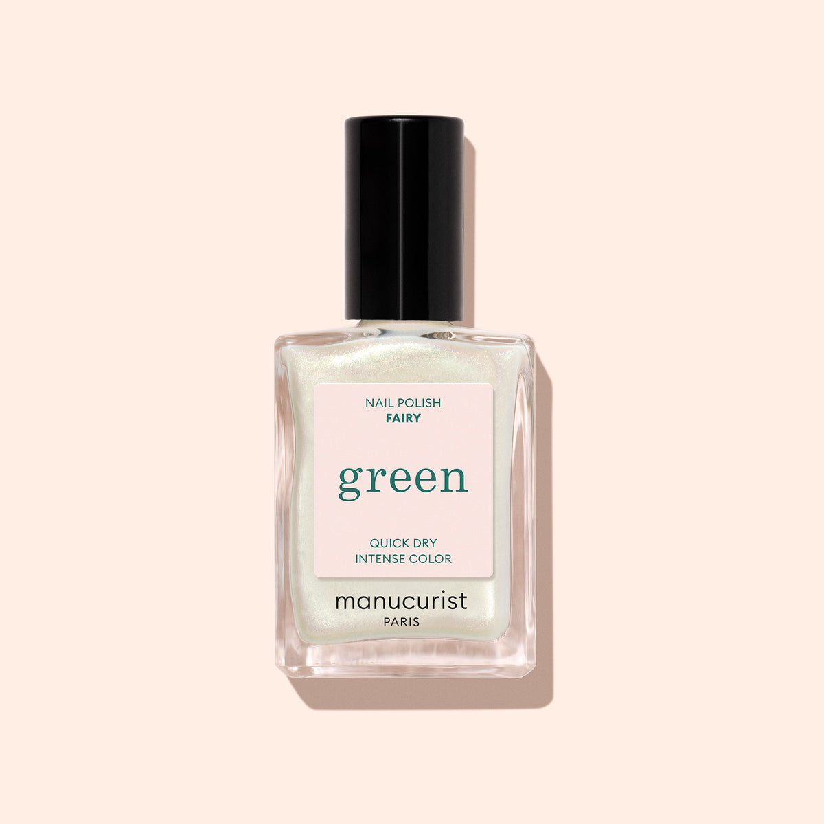 A clear glass bottle of Manucurist Fairy nail polish with a black cap, featuring green sparkle and labeled QUICK DRY and INTENSE COLOR, is set against a pale peach background.