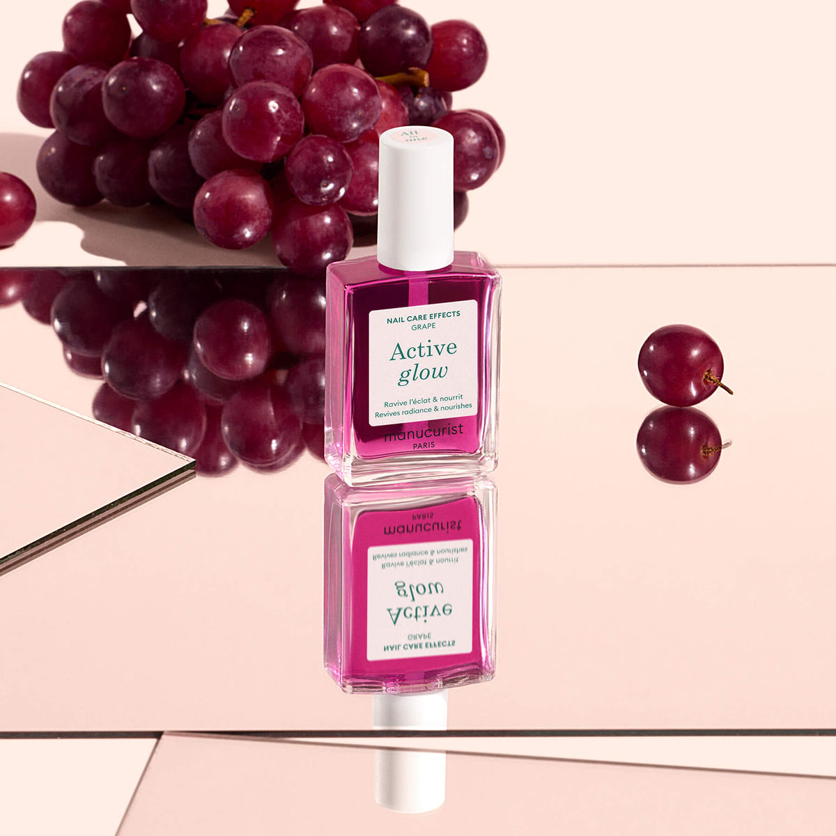 Un flacon de soin pour ongles Active™ Glow Grape de Manucurist posé sur une surface miroitante, avec des grains de raisin en arrière-plan et son reflet visible en dessous, offrant une brillance naturelle à vos ongles.