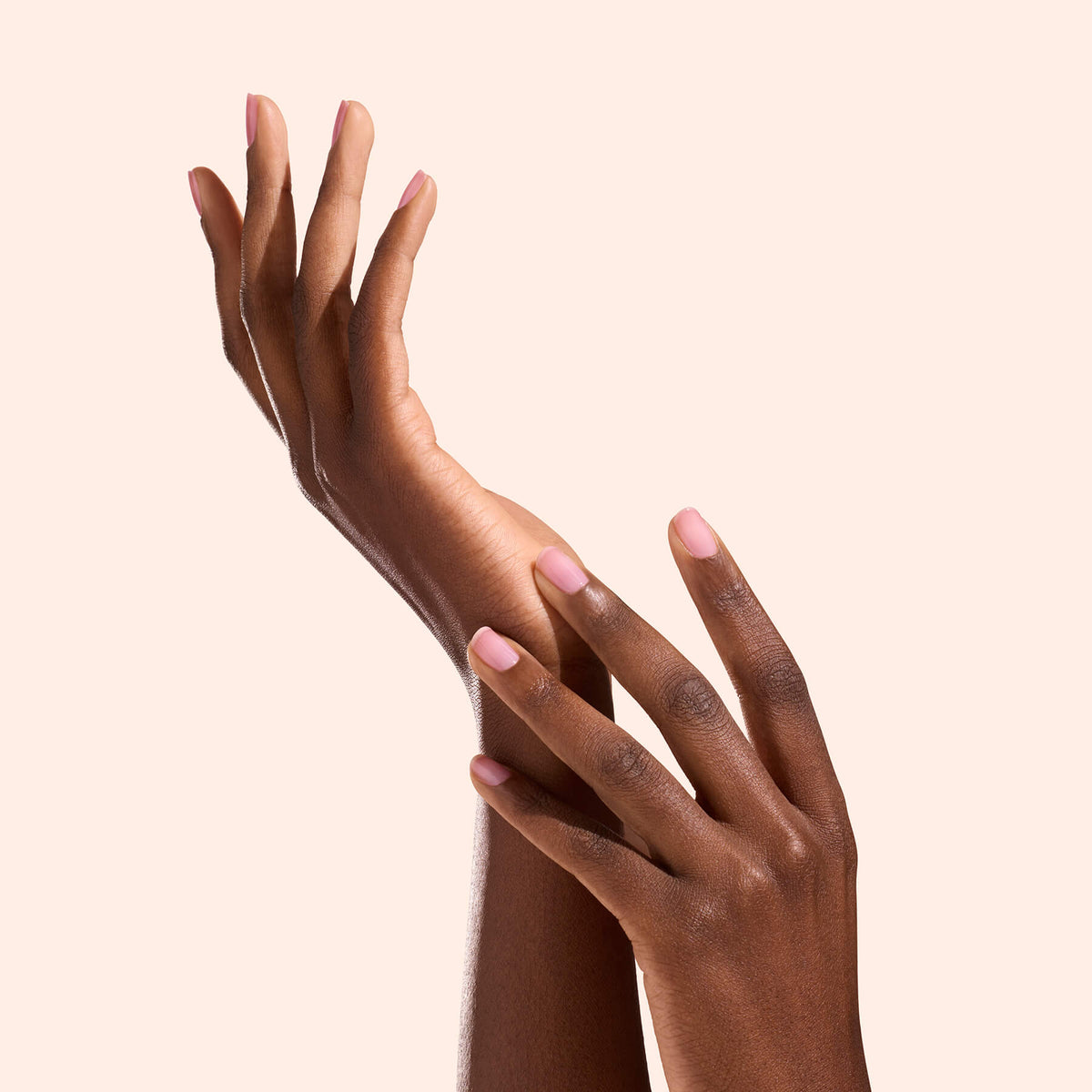 Deux mains à la peau foncée, les doigts légèrement écartés et les ongles soigneusement manucurés avec la brillance naturelle d’Active™ Glow Grape de Manucurist, se touchent délicatement sur un fond beige clair uni.