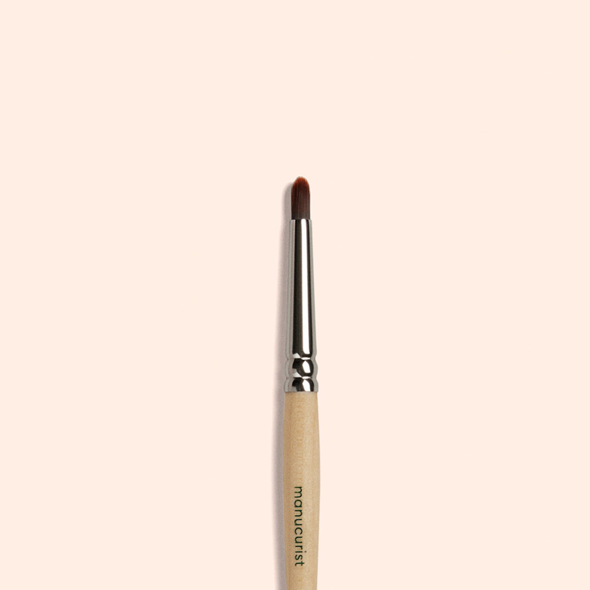 Manucurist’s Glitter Brush—a small brush with a wooden handle, metal ferrule, and soft vegan bristles—sits centered on a light beige background. The handle features "manucurist" branding, perfect for adding sparkle to your manicure.