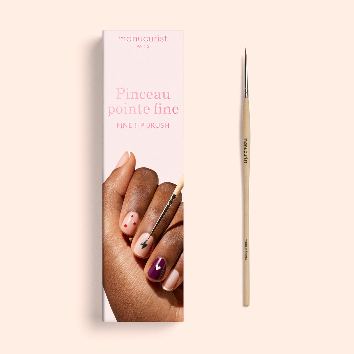 A Fine Tip Brush by Manucurist with vegan synthetic bristles is placed next to its box, which features images of nails painted in multiple colors and designs. The packaging displays the text “manucurist PARIS” and “Fine Tip Brush / Pinceau pointe fine.”.