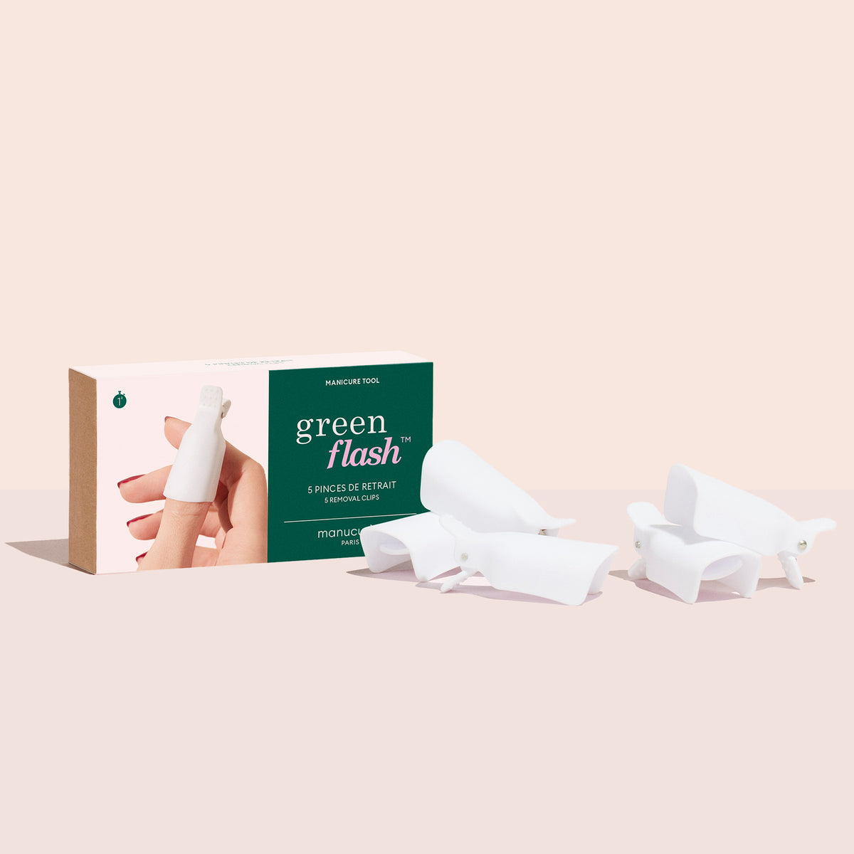 A box labeled Green Flash™ Remover Clips Duo by Manucurist-us is shown with a hand wearing a white nail cap. Five white reusable clips for gel polish removal are displayed on a light surface beside the box.