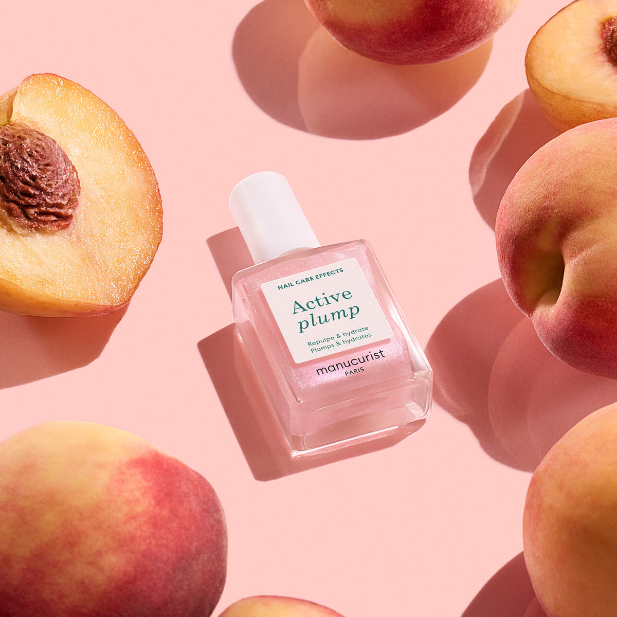 Un flacon transparent de Manucurist Active™ Plump Pink Glazed repose sur une surface rose, entouré de pêches entières et coupées en deux, créant une esthétique fraîche et vibrante qui met en valeur les bienfaits d&