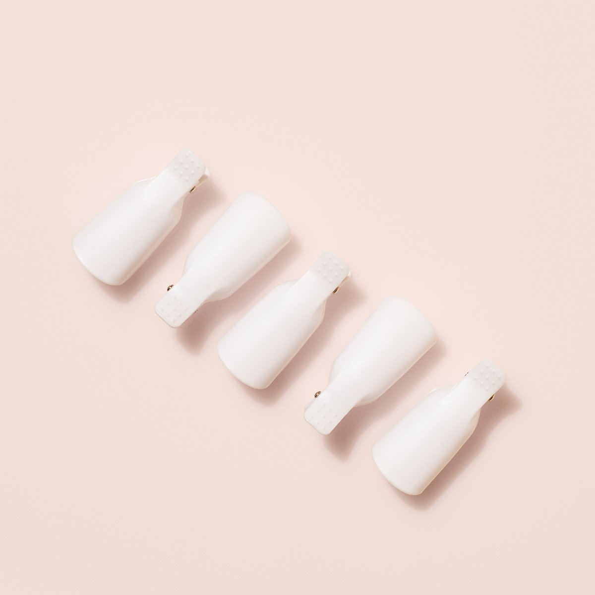 Five white plastic nail clips from Manucurist-us Green Flash™ Remover Clips Duo are arranged in a neat diagonal line on a light beige background. Perfect for use with plant-based nail polish remover.