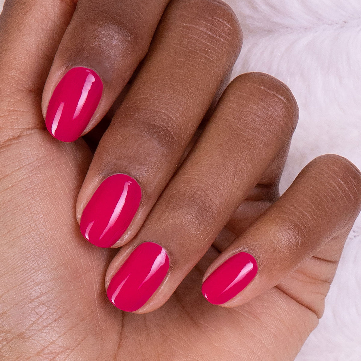 A hand with medium-dark skin tone shows neatly manicured nails painted in glossy "Fuchsia" gel polish by Manucurist, set against a soft white background.