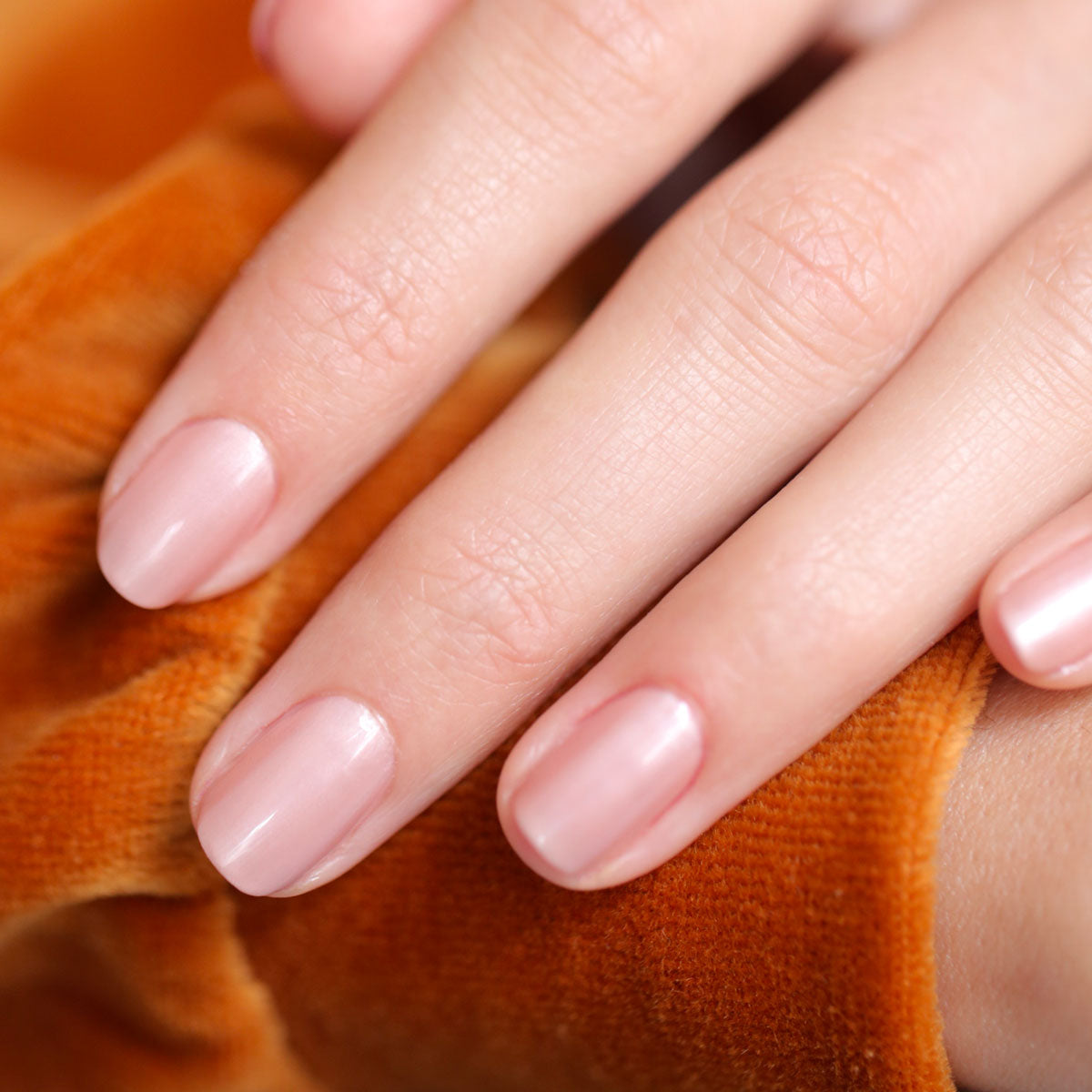 A close-up of a hand with neatly manicured nails painted in Carnation by Manucurist rests on orange velvet fabric.