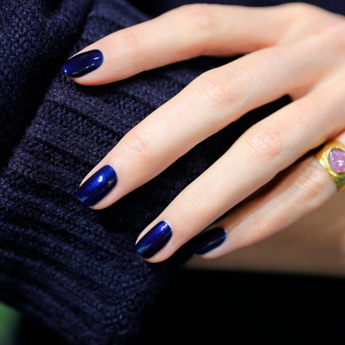Une main aux ongles impeccablement manucurés avec le vernis "Dark Night" de Manucurist repose sur une maille bleu foncé, ornée d&