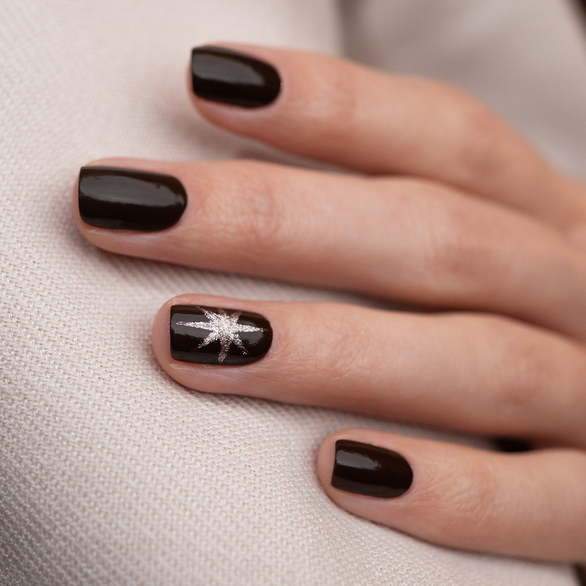 A hand with short, well-groomed nails painted in Manucurist’s Clove gel polish, a glossy chocolate brown. The ring finger showcases a silver starburst design. The hand is resting on light fabric.