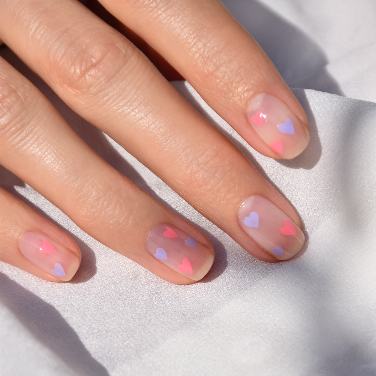 A hand with natural nails features small pink and purple heart designs created using the Manucurist Dotting Tool Nail Art Liner Brush Duo, shown close-up against a white fabric background.