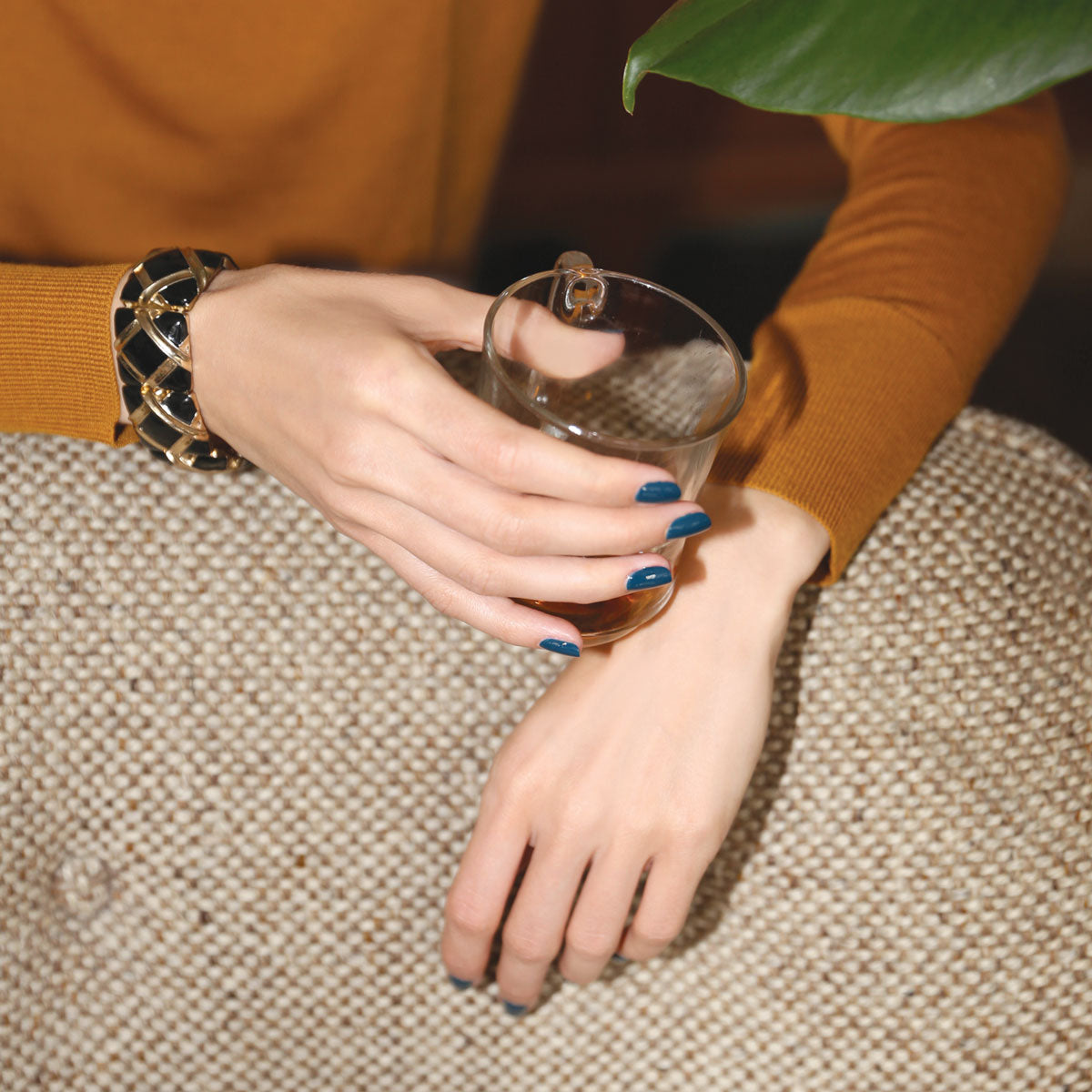 Portant la teinte Dark Clover de Manucurist, une personne aux ongles bleu canard tient un verre contenant un liquide ambré, vêtue d&