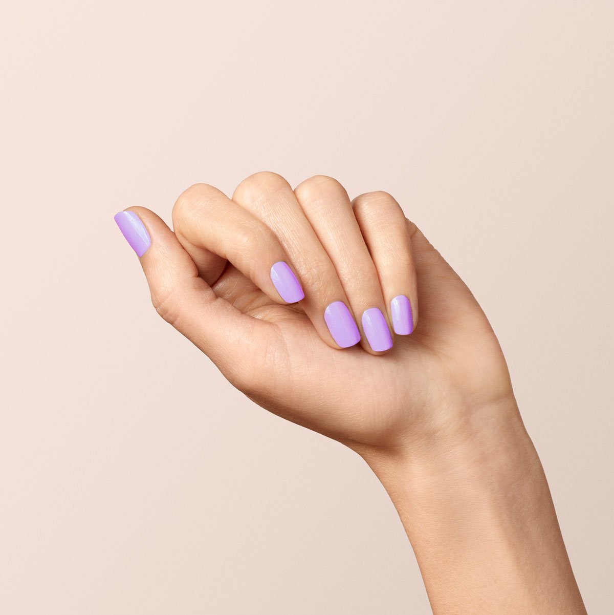 Une main à la peau claire, sur un fond neutre, présente des ongles soigneusement manucurés et peints avec Lisa Lilas de Manucurist, un vernis gel lilas brillant qui apporte une touche de couleur fraîche et de subtiles vibes des années 90.