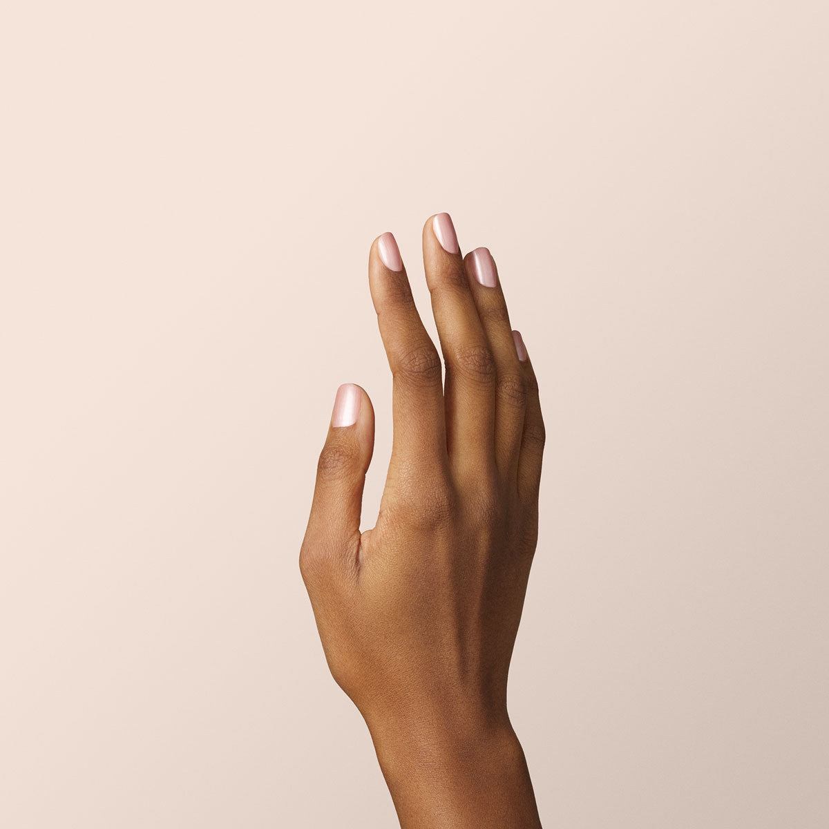 A hand with medium brown skin is held upright against a light beige background, fingers slightly spread and neatly trimmed natural nails—perfectly prepped for a Manucurist Carnation Green Flash gel polish manicure.