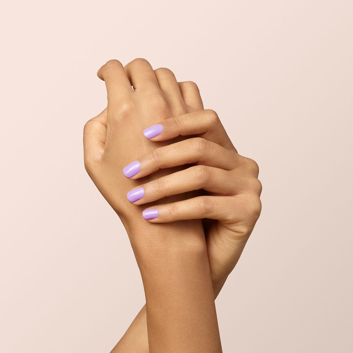 Deux mains se tiennent délicatement sur un fond clair, leurs ongles soigneusement manucurés et vernis avec Lisa Lilas de Manucurist, un vernis gel lilas brillant qui apporte de subtiles vibes des années 90 à ce look élégant.