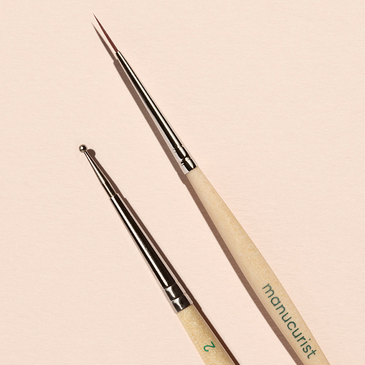 The Manucurist Dotting Tool Nail Art Liner Brush Duo features light wooden handles and silver tips, and is shown arranged on a pale pink background.