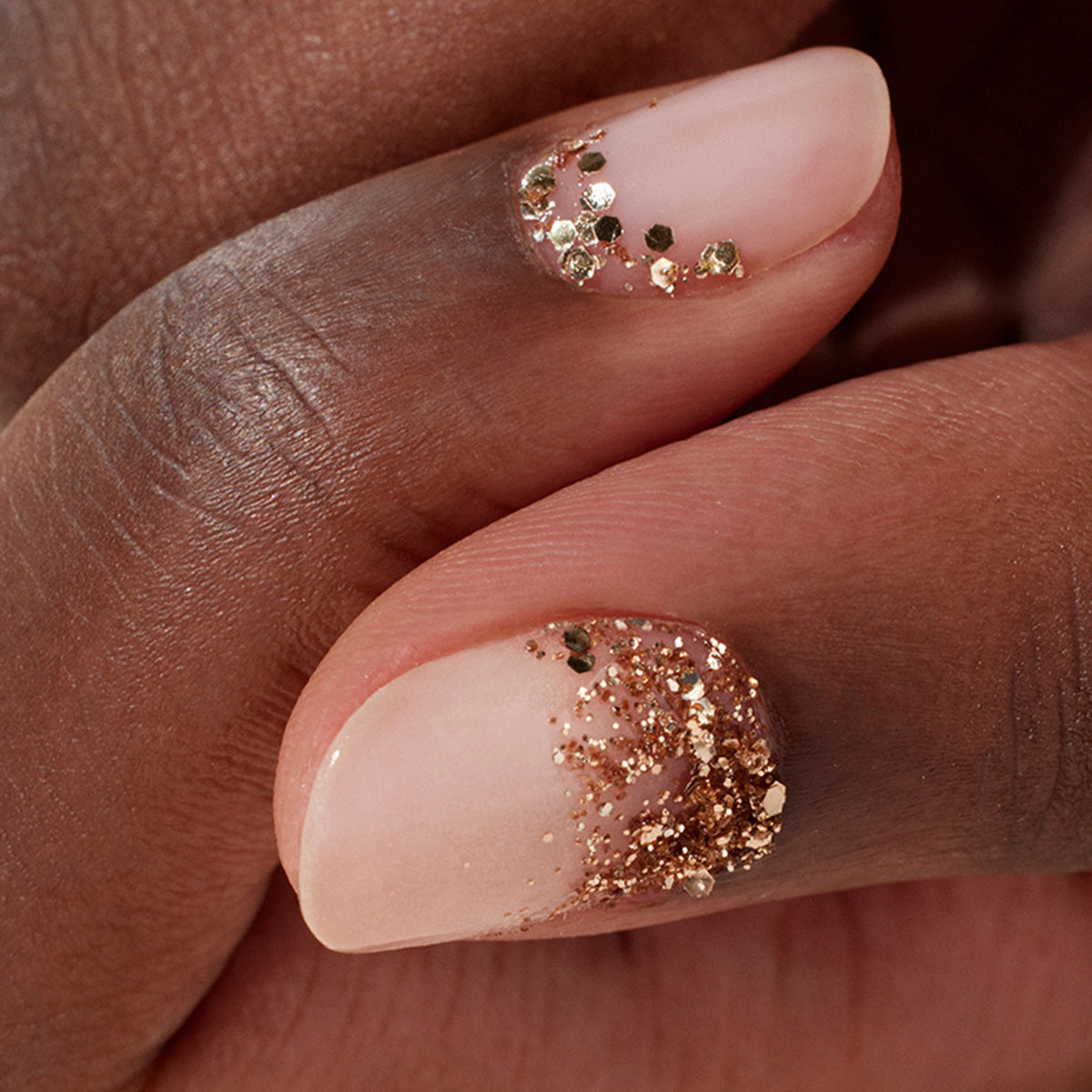 Deux doigts aux ongles nude rehaussés de Paillettes Biodégradables Rose Gold écoresponsables de Manucurist sur les cuticules, créant un Nail Art chic et élégant.