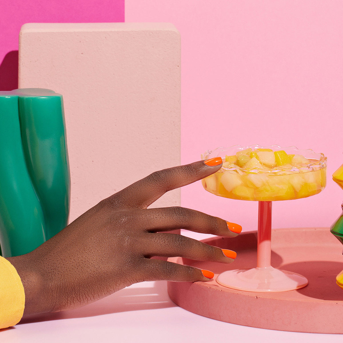 Une main avec le vernis à ongles orange vif Sunset de Manucurist attrape un plat en verre rose de salade de fruits, posé parmi des objets géométriques colorés sur un fond rose, pour une ambiance moderne et dynamique.