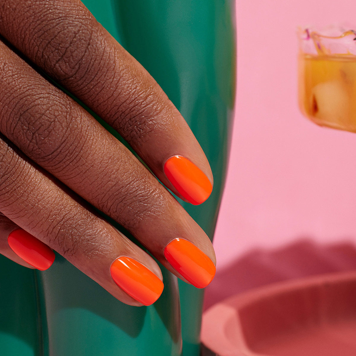 Une main aux ongles soigneusement manucurés et vernis avec Sunset de Manucurist, un vernis orange vif, repose sur une surface verte brillante, avec un fond rose et une boisson avec des glaçons partiellement visible.