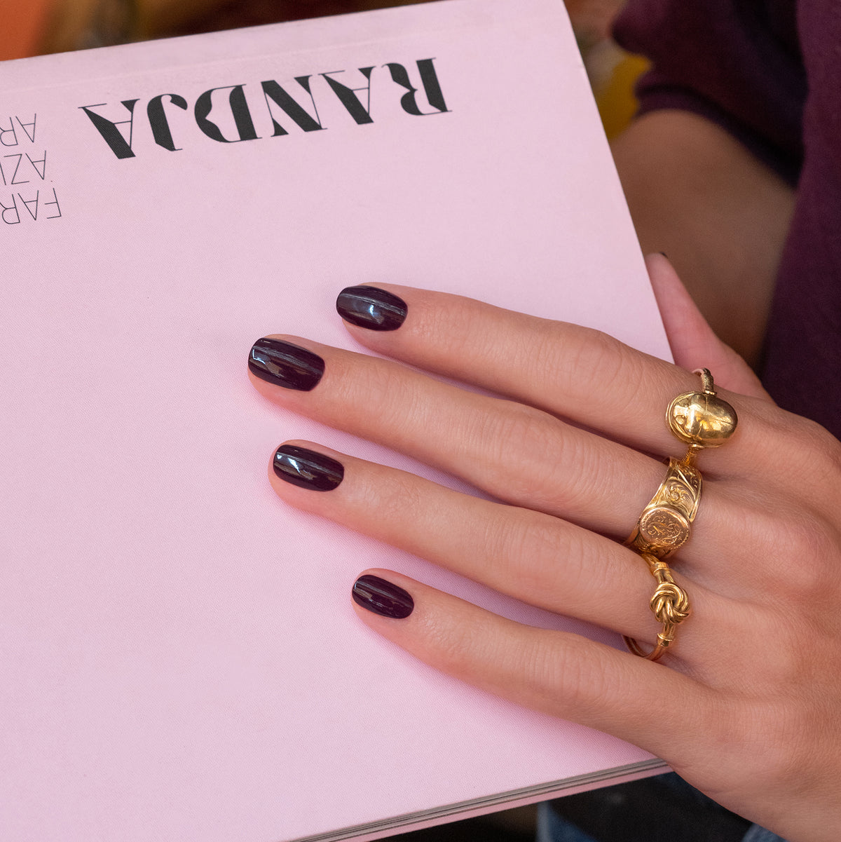 A hand with Manucurist’s Aubergine gel polish and gold rings holds a light pink book with RANDA written upside down on the cover.