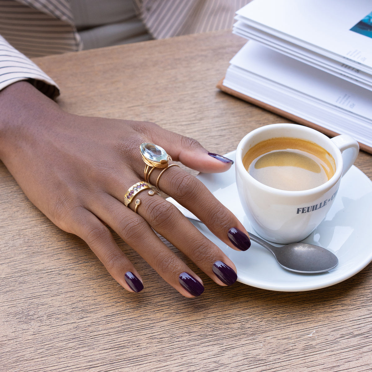 A hand with Manucurist’s Aubergine gel polish and multiple gold rings rests on a wooden table next to an espresso, a spoon, and an open notebook.