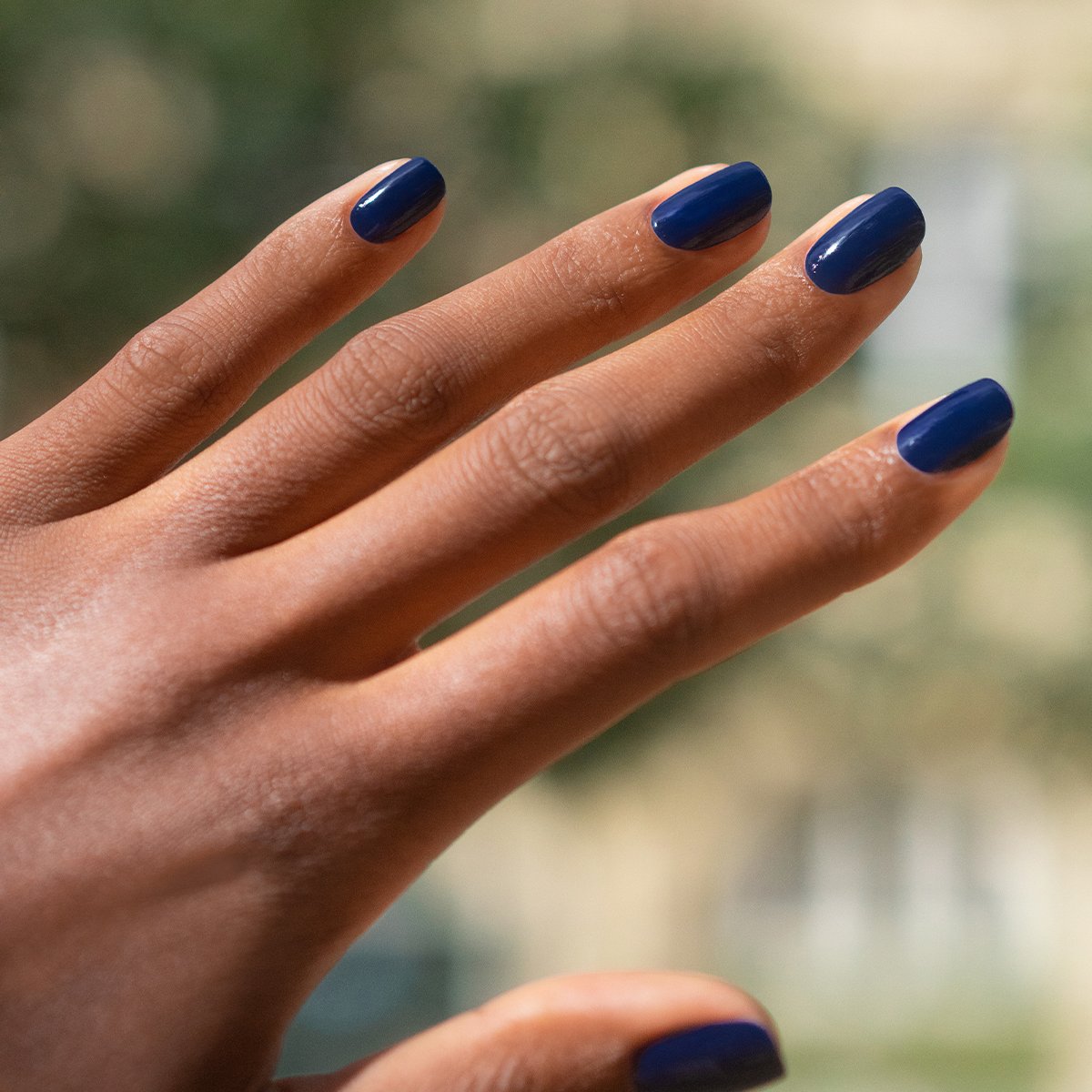 A hand with dark skin and neatly manicured nails is shown, each nail painted in Manucurist’s Navy Blue gel polish, against a softly blurred outdoor background.