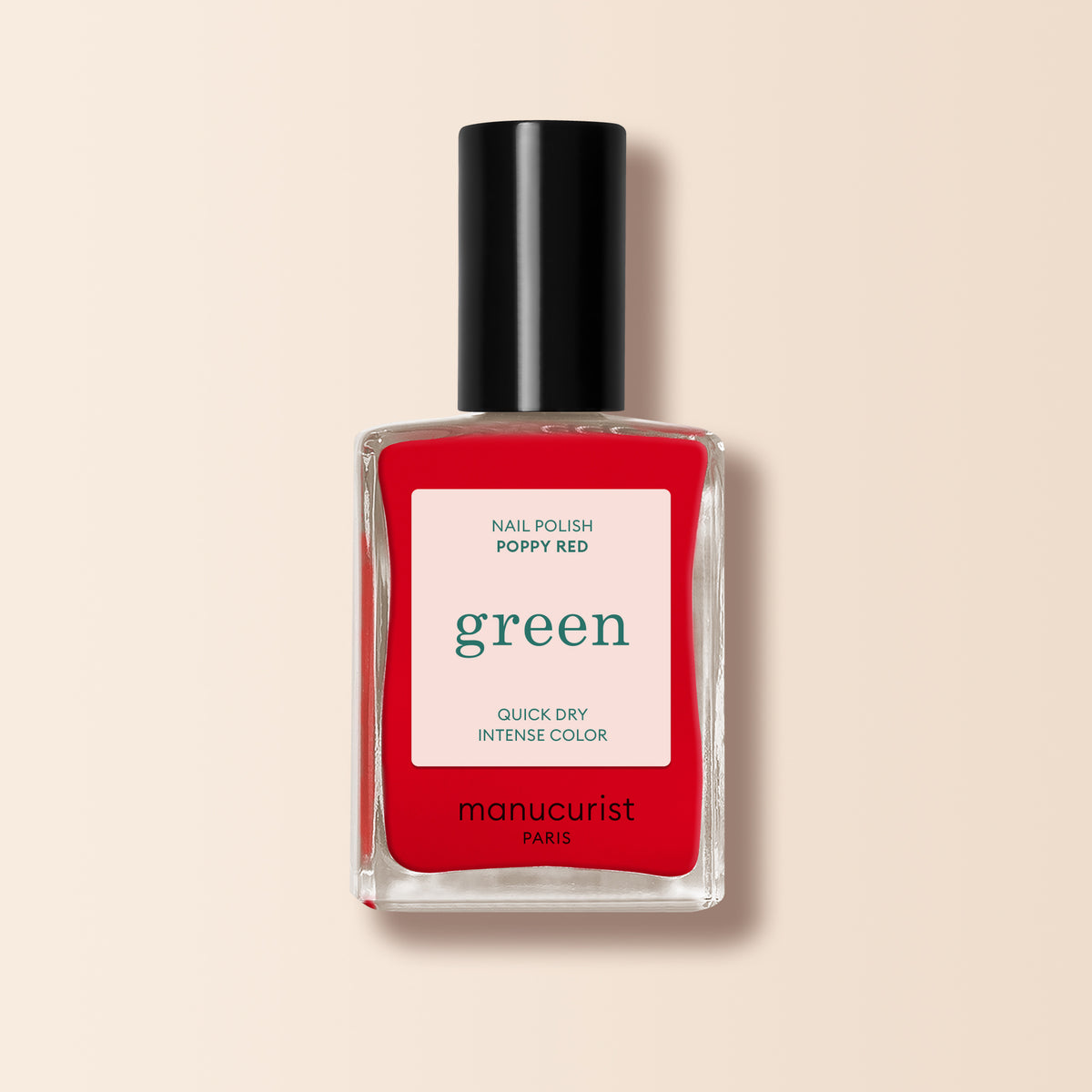 A clear glass bottle of Manucurist Poppy Red nail polish features a black cap and white label stating "quick dry intense color." The vibrant orangey red shade stands out against a soft beige background.