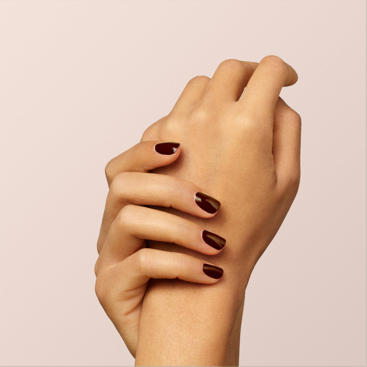 Two hands with neatly manicured nails painted in Clove, a rich chocolate brown from Manucurist, are gently clasped together against a soft, light beige background.