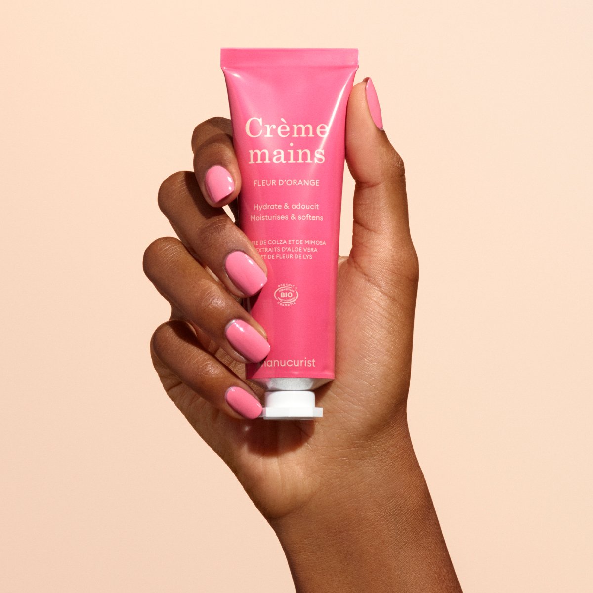 Une main avec du vernis à ongles rose tient un tube rose de crème pour les mains Manucurist Everyday Orange Blossom - Cosmos Organic, enrichie en huile de coco, sur un fond beige.