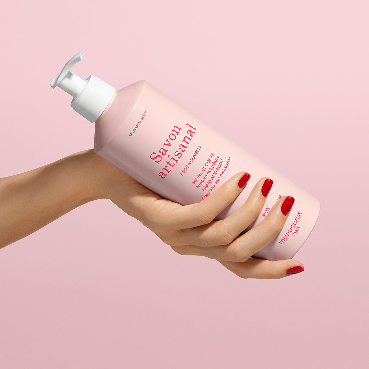 A hand with red nail polish holds a pink bottle of Manucurist Organic Rose Hand Body Wash - Cosmos Organic with a white pump, set against a matching pink background.