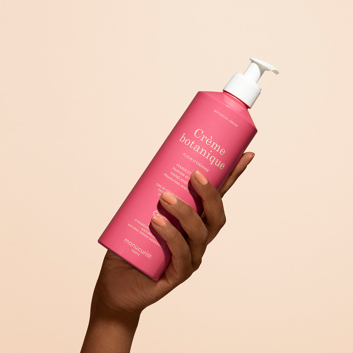 A hand holds a pink bottle of Manucurist Soothing Orange Blossom Hand Body Cream - Cosmos Organic with a white pump dispenser against a plain beige background.