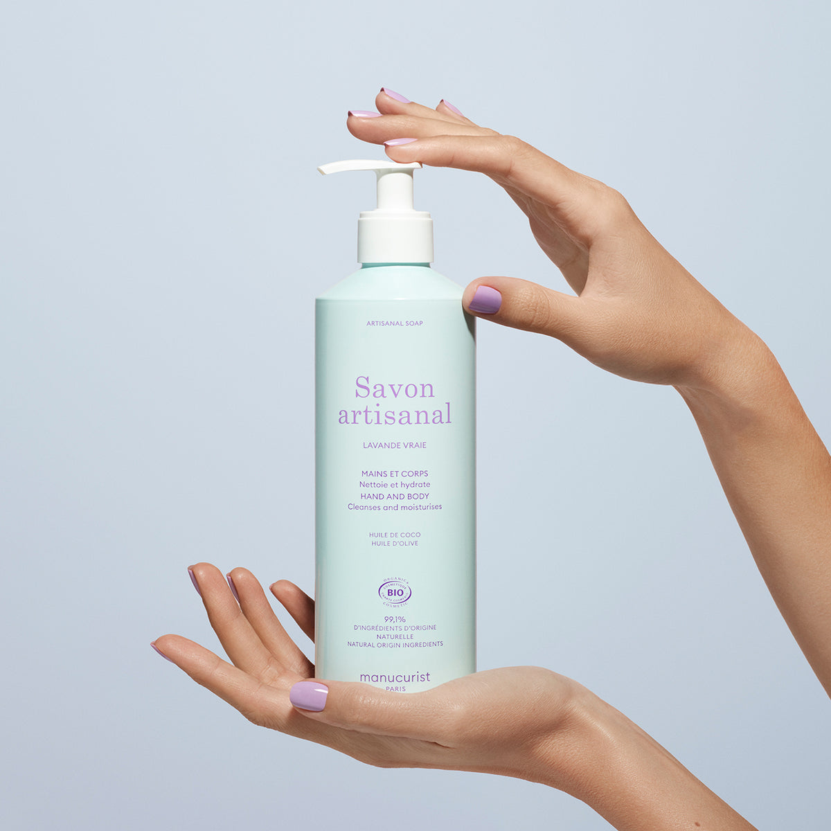 A person with lavender-painted nails holds a light blue pump bottle of Manucurist’s Organic Lavender Hand Body Wash - Cosmos Organic against a light blue background, suggesting a soothing lavender scent.