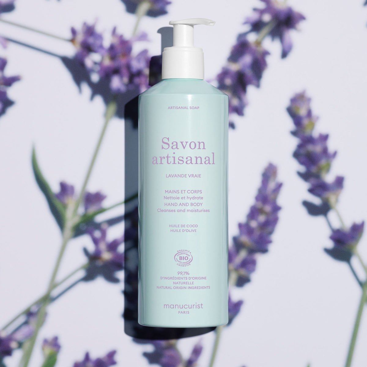 A light blue bottle of Manucurist Organic Lavender Hand Body Wash - Cosmos Organic with a pump sits on a surface with lavender flowers and soft shadows. The label highlights coconut and olive oil, natural ingredients, and a subtle lavender scent.
