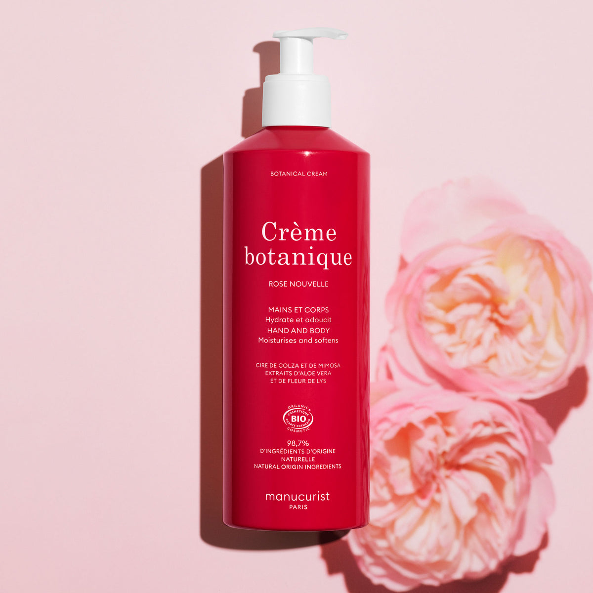 A pink bottle of Manucurist Soothing Rose Hand Body Cream - Cosmos Organic stands upright on a light pink background beside two pink roses. The white pump and text highlight its promise to deeply hydrate your skin.