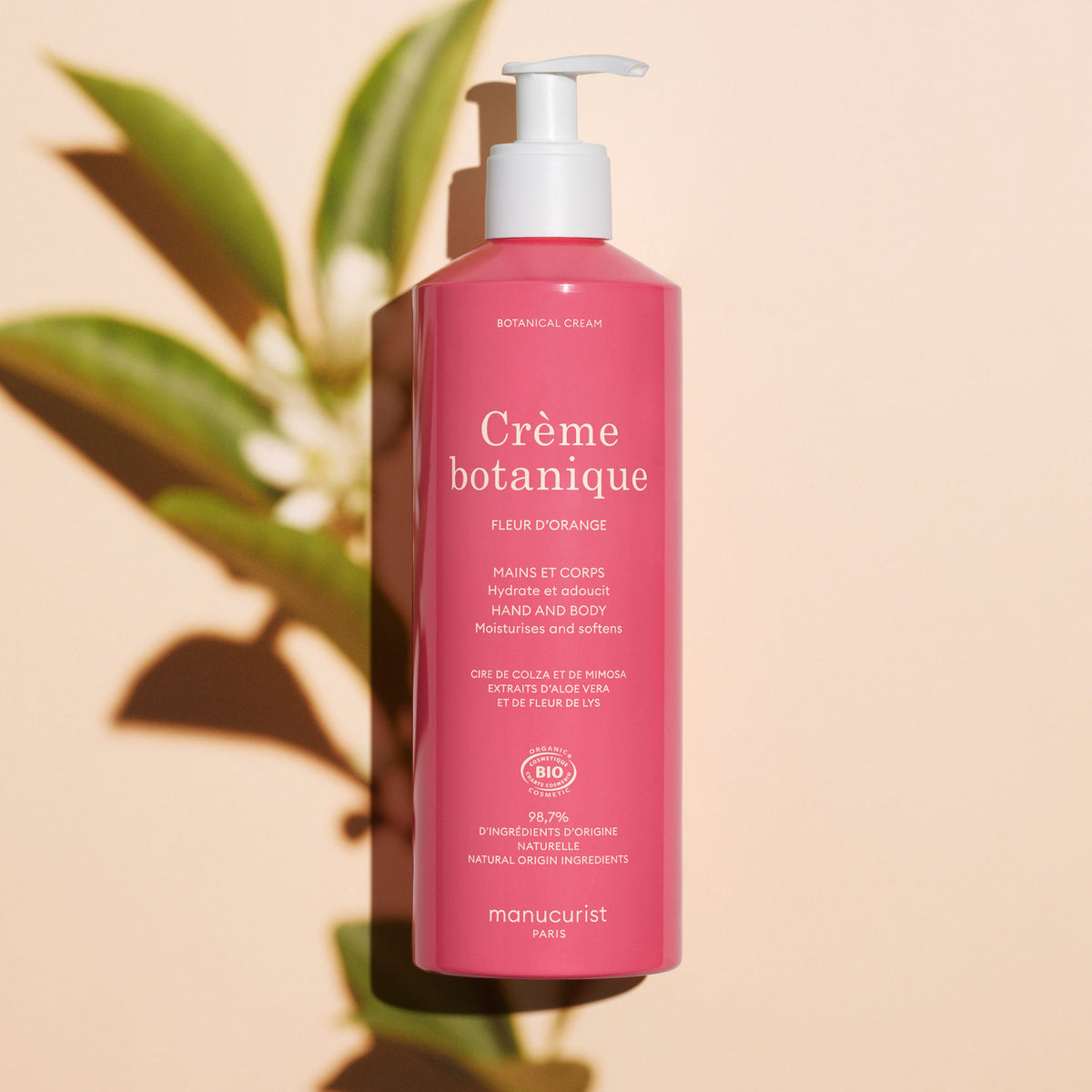 A pink bottle of Manucurist Soothing Orange Blossom Hand Body Cream – Cosmos Organic with a pump dispenser stands on a light surface, casting a green leafy shadow. The bilingual label highlights its orange blossom fragrance.