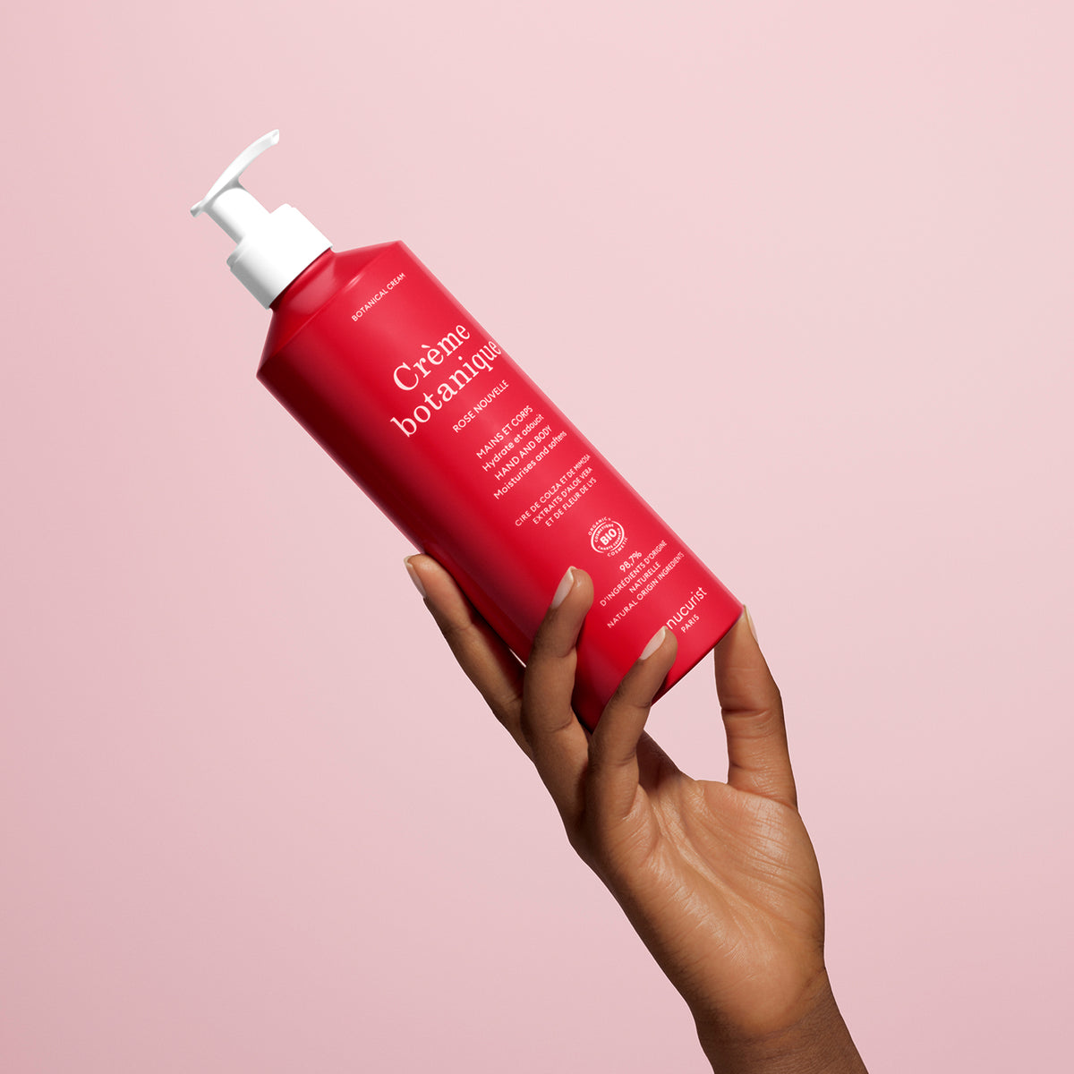 A hand with dark skin holds up a bright red Manucurist Soothing Rose Hand Body Cream - Cosmos Organic with a white pump, against a pale pink background. The label features white text, and the product is designed to deeply hydrate skin.