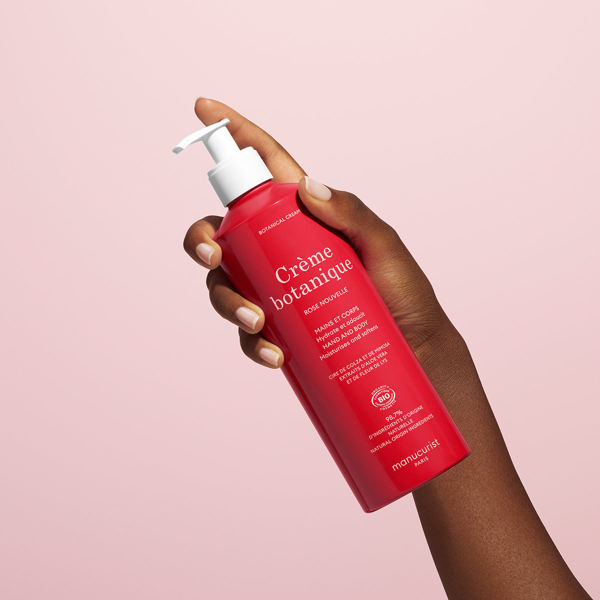 A hand holds a red bottle of Manucurist Soothing Rose Hand Body Cream - Cosmos Organic with a white pump, against a light pink background. The label features white text highlighting its natural origin and deep hydration benefits.