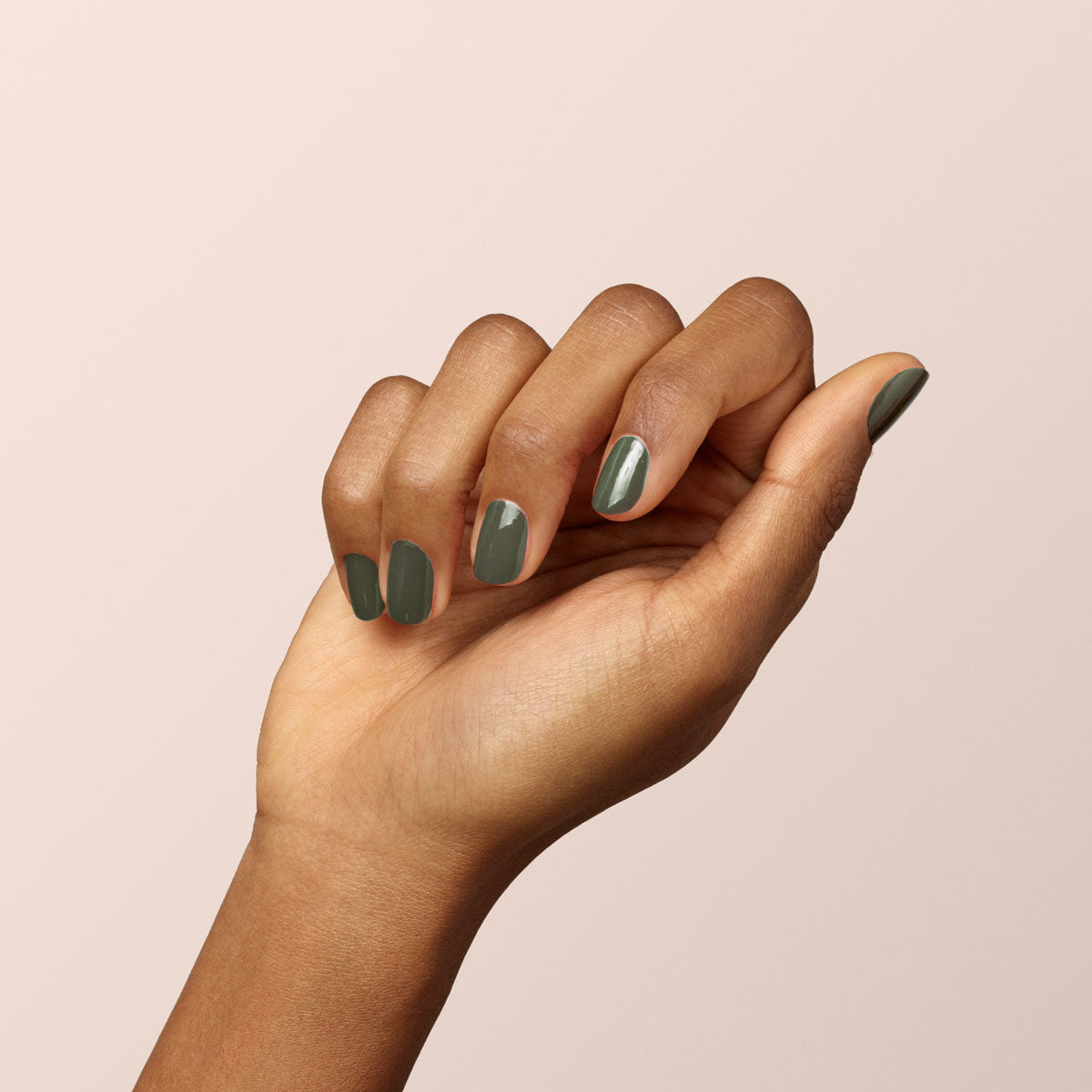 A hand with medium-brown skin is held upright against a light beige background. The nails are painted with Manucurist’s Khaki, a glossy olive green polish featuring a subtle grey undertone.