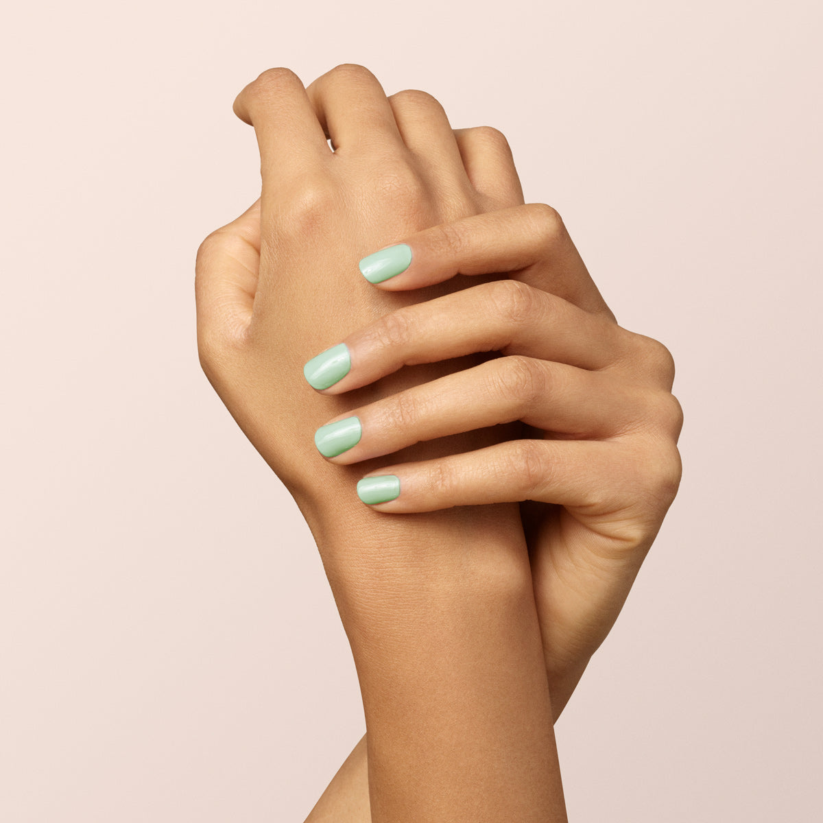 A person’s hands with interlaced fingers, showing neatly manicured nails painted with Manucurist Mint pastel green nail polish, against a plain light beige background.