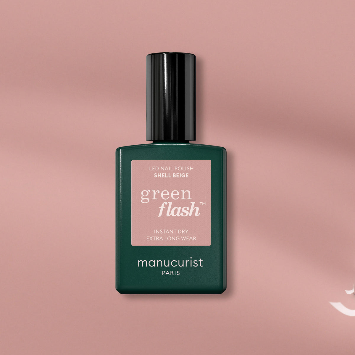 A bottle of Manucurist Shell Beige, a pinkish beige gel polish, stands on a pink background with a soft shadow. The dark green bottle features a pale pink label.