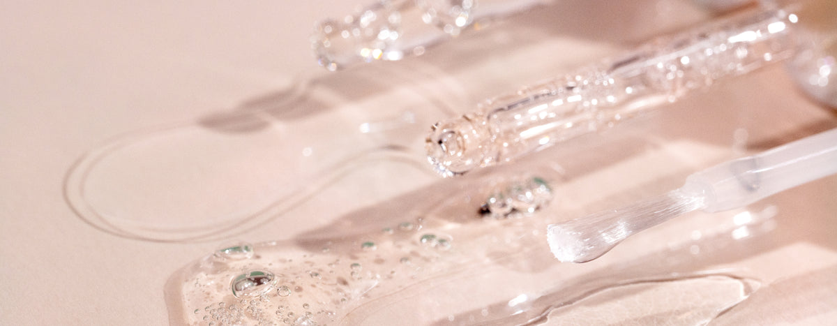 Close-up of clear glass droppers and a brush applicator with transparent gel serum and bubbles spilled on a light, reflective surface. The scene is softly lit with a pinkish hue.