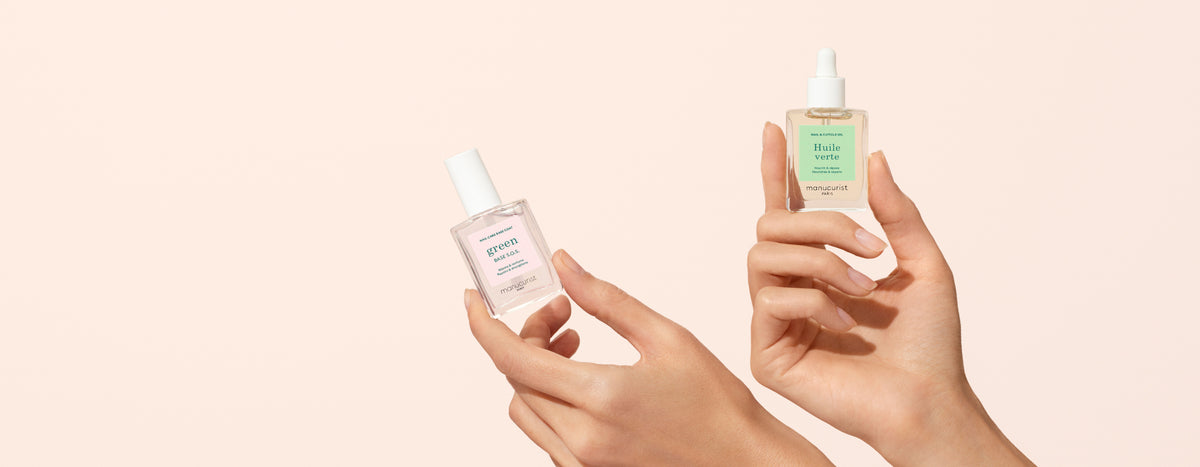 Two hands hold up glass dropper bottles of skincare serum against a light beige background. The left bottle has a white label, and the right bottle has a green label and a dropper cap.