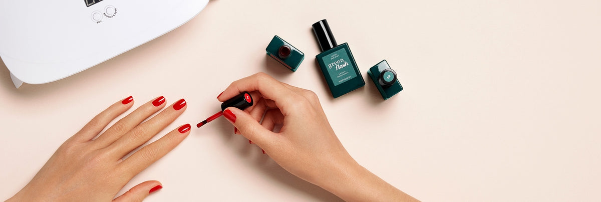 A person paints their fingernails with red nail polish, using their right hand. Three bottles of nail polish and a white UV lamp are placed nearby on a light-colored surface.