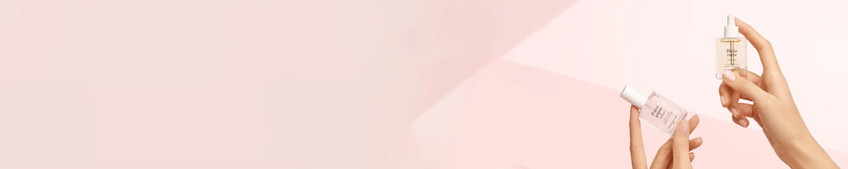 Two hands hold glass skincare serum bottles against a soft pink background; one bottle is clear with a white cap, and the other is tinted with a dropper lid.